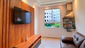 A modern living room with a flat-screen TV mounted on a wooden wall, a brown leather sofa, a built-in fireplace, and a window with a view of buildings and greenery.