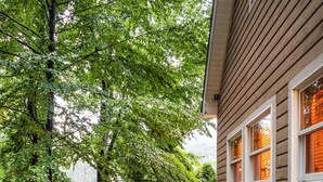 A wooden deck leading to a house with a covered porch, surrounded by trees and a stack of firewood.