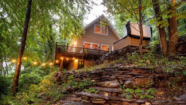 A rustic cabin with a stone staircase leading to a wooden deck, surrounded by lush greenery.