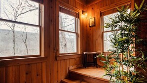 A wooden cabin interior with a staircase leading to a windowed loft area, a single chair, and a potted plant.