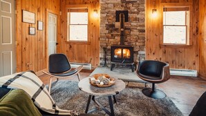 A cozy cabin interior with wooden walls, a stone fireplace, two modern chairs, and a small round table with decorative items.