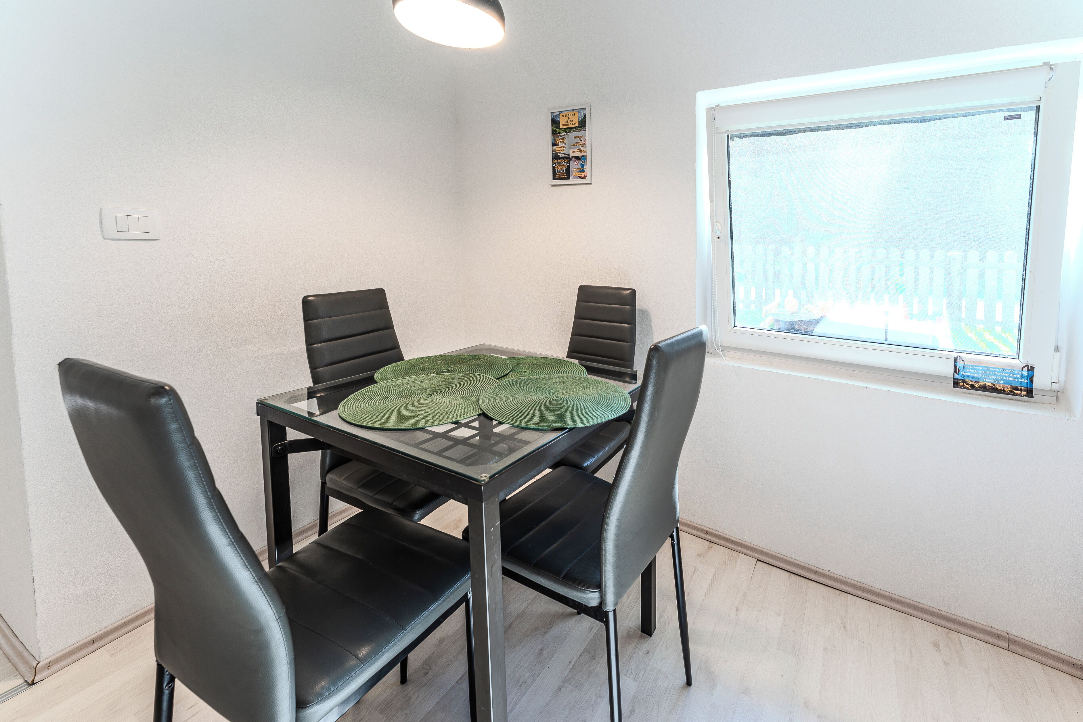 A dining area with a glass table, four black chairs, and a window with a view of a fence.