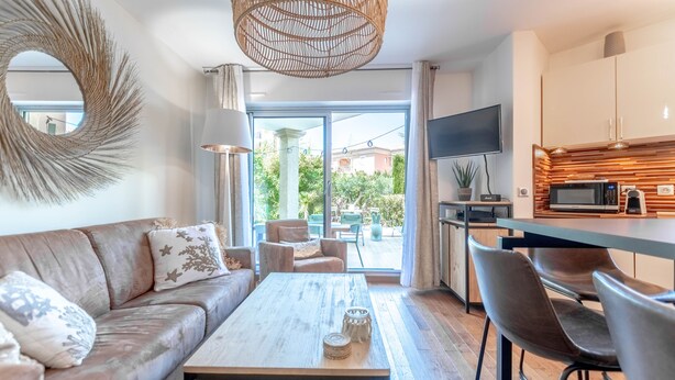 A modern living room with a sofa, coffee table, and chairs, a kitchen with a TV and microwave, and a large window with a view of greenery.