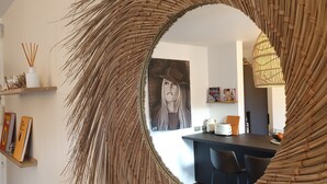 A room with a large woven mirror, a painting on the wall, and a kitchen area with chairs and a counter.