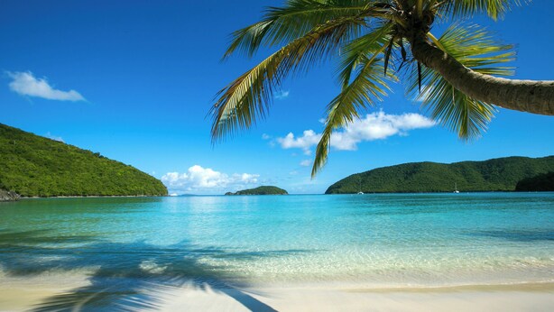 Ein Strand mit klarem türkisblauem Wasser, weißem Sand und grünen Hügeln im Hintergrund.