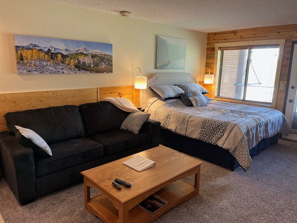 A cozy room with a bed, a sofa, and a coffee table. There are two paintings on the wall, a lamp, and a window with blinds.