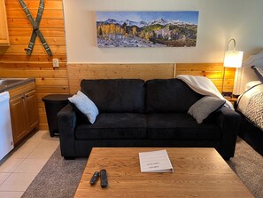 A cozy room with a wooden floor, a black sofa, a wooden coffee table, and a painting of a mountain landscape.