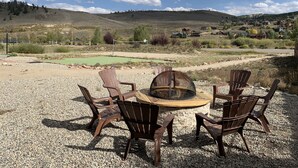 Una mesa redonda de madera con sillas y un hogar a leña en un área de grava, con vista a un campo de golf y casas al fondo.