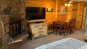 Una habitación acogedora con chimenea de piedra, un televisor sobre un mueble de madera, una mesa de comedor de madera con sillas, y una colcha de cama estampada.