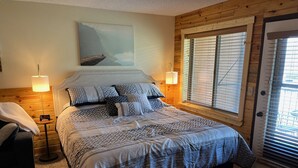Un dormitorio con una cama grande, dos lámparas de noche, un cuadro en la pared y una ventana con persianas.