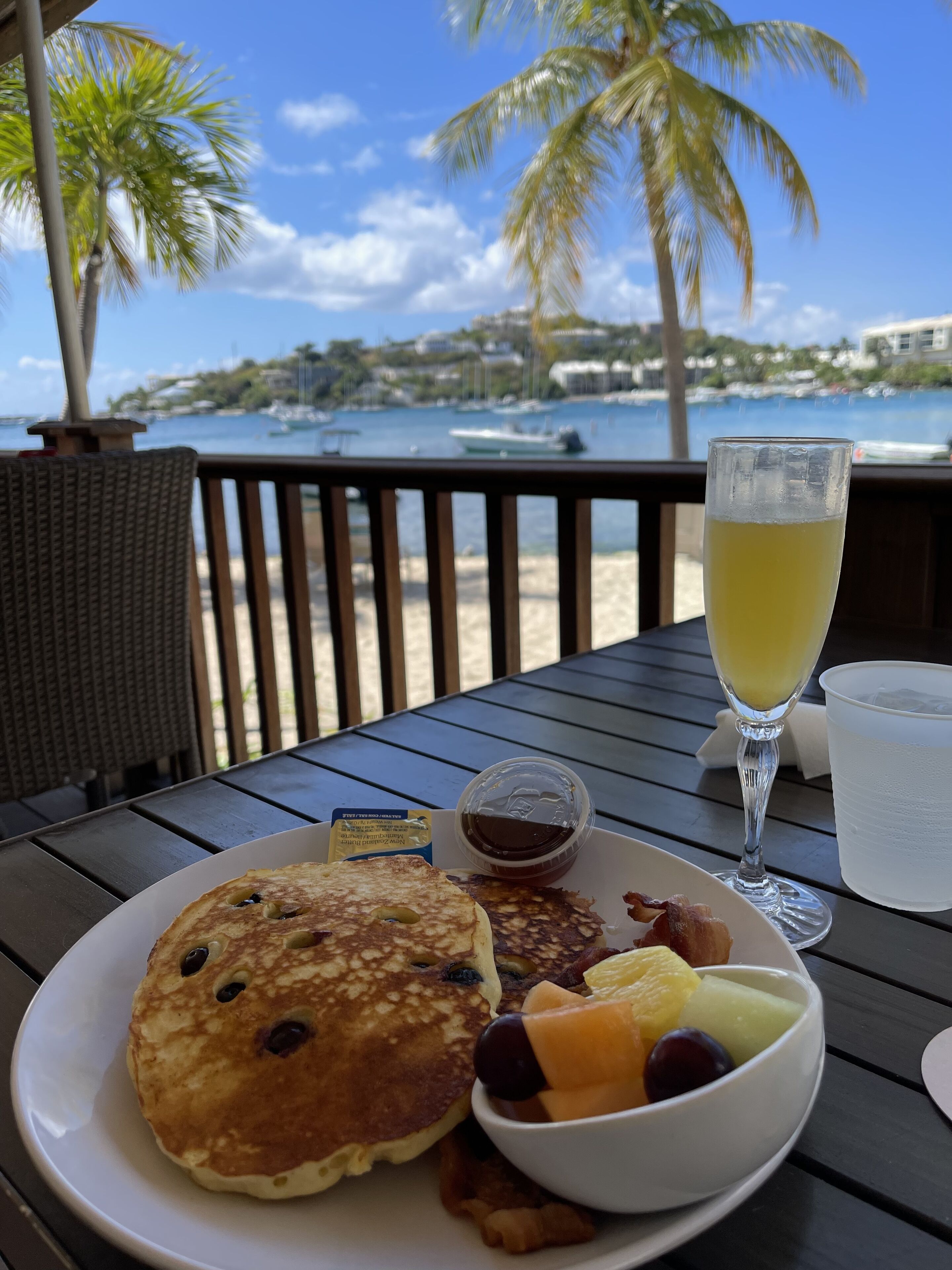 A breakfast plate with pancakes, bacon, and fruit, a glass of orange juice, and a small container of syrup, set on a table with a view of the beach and palm trees.