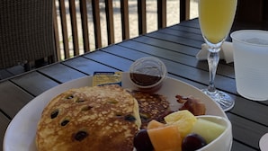 Une assiette de petit-déjeuner avec des crêpes, du bacon et des fruits, un verre de jus d’orange et un petit récipient de sirop, le tout posé sur une table offrant une vue sur la plage et les palmiers.