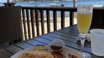 A breakfast plate with pancakes, bacon, and fruit, a glass of orange juice, and a small container of syrup, set on a table with a view of the beach and palm trees.