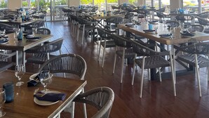 Un restaurant spacieux avec de hauts plafonds, des poutres apparentes et de grandes fenêtres offrant une vue sur l’extérieur. Les tables sont accompagnées de chaises, et l’intérieur est agrémenté de luminaires suspendus et de ventilateurs de plafond.
