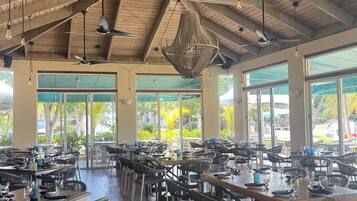 A spacious restaurant with high ceilings, exposed wooden beams, and large windows offering views of the outdoors. Tables are set with chairs, and the interior features hanging light fixtures and ceiling fans.