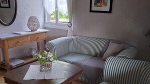 A living room with a striped sofa, a wooden coffee table with a vase of flowers, a desk with a lamp, and a window with curtains. - Quiet 1st floor apartment with balcony and patio, 18 km from the sea (Landebia frankreich)