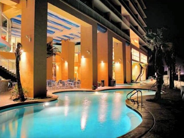 An indoor swimming pool with a curved design, surrounded by palm trees and a covered area with seating. - Splash Kids Paradise 17th floor is clean, spacious plus free daily beach service (Panama City Beach)