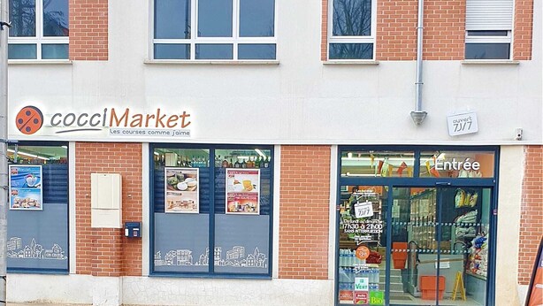 Een gebouw met twee verdiepingen met een etalage genaamd 'cocciMarket', voorzien van een stadsgezichtsmural, een glazen ingang en een bord waarop 'ouvert 7J/7' staat.