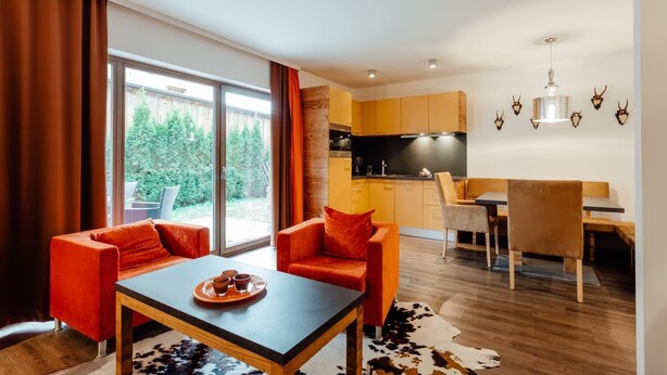 Een moderne woonkamer met een bank en een eettafel, een keuken met gele kastjes en een groot raam met gordijnen.