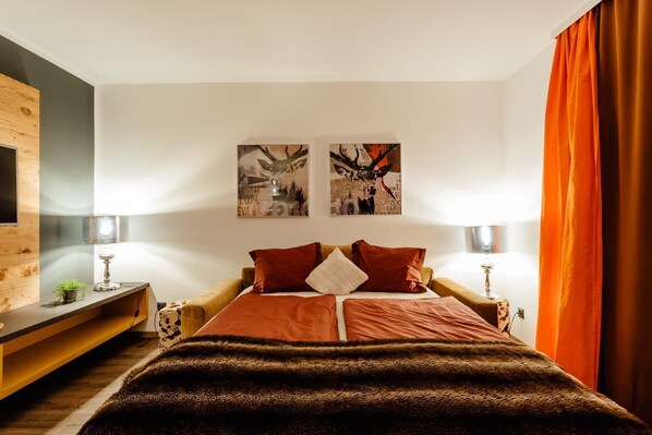 A modern bedroom with a wooden headboard, two wall-mounted lamps, a large bed with a brown blanket, and two framed pictures above the bed.