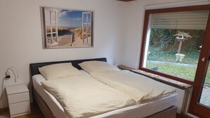 Een slaapkamer met een houten bed, een nachtkastje, een stopcontact en een raam met uitzicht op een tuin.