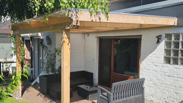 Een terras met een houten pergola, buitenmeubels en een grote schuifdeur naar een overdekte ruimte.