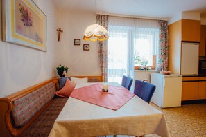 A dining area with a table, chairs, a bench, a chandelier, a refrigerator, and a window with curtains.