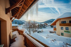 A snow-covered balcony with wooden railings and a view of a snowy village and mountains.