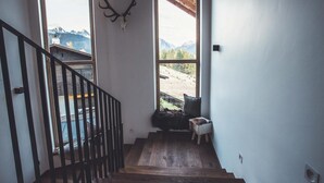 Uma escadaria com degraus de madeira leva a uma varanda, um crânio de veado montado na parede, e vista para montanhas através de grandes janelas.