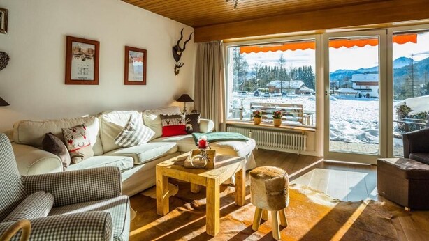 Ein gemütliches Wohnzimmer mit Holzbalkendecke, einer Sofagarnitur, einem Couchtisch und Blick auf eine verschneite Landschaft durch große Fenster.