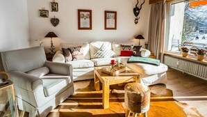 Ein gemütliches Wohnzimmer mit einem karierten Sofa, einem Holztisch und einem Fellstuhl. An den Wänden hängen Bilderrahmen und das Fenster bietet Blick ins Freie.