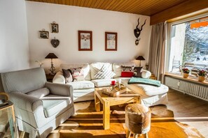 A cozy living room with a checkered sofa, a wooden coffee table, and a fur chair. There are framed pictures on the wall and a window with a view of the outdoors.