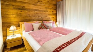 Ein Doppelbett mit rot-weiß karierter Tagesdecke und roter Bordüre. Ein hölzerner Kopfseitenborden und ein Nachttisch mit Lampe sind ebenfalls vorhanden. Der Raum verfügt über Holzvertäfelung an den Wänden und ein Fenster mit Vorhängen.