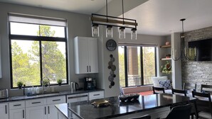 Eine moderne Küche mit einer großen Kochinsel, weißen Schränken, einer Kaffeemaschine und Blick auf Bäume durch große Fenster.