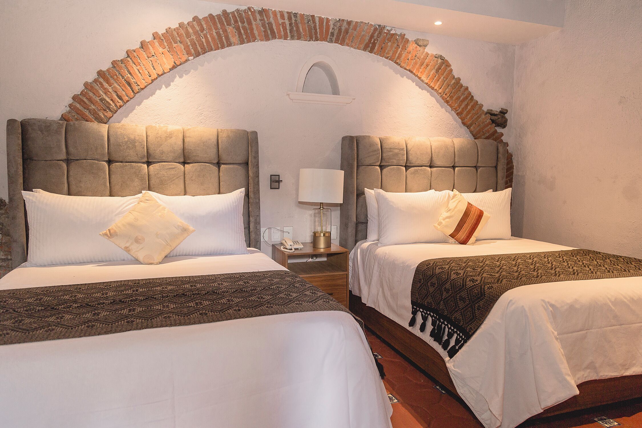 A hotel room with two beds, each with a headboard, a nightstand, a lamp, and a brick arch above the beds.