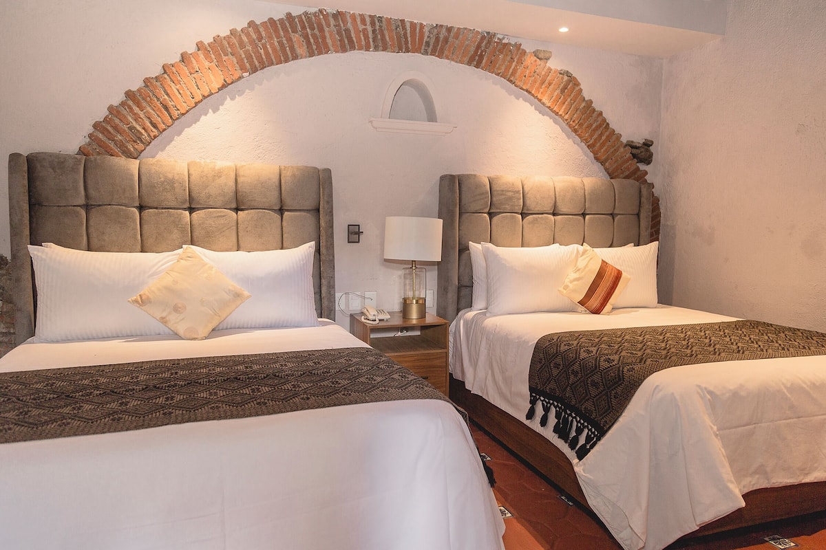 A hotel room with two beds, each with a headboard, a nightstand, a lamp, and a brick arch above the beds.