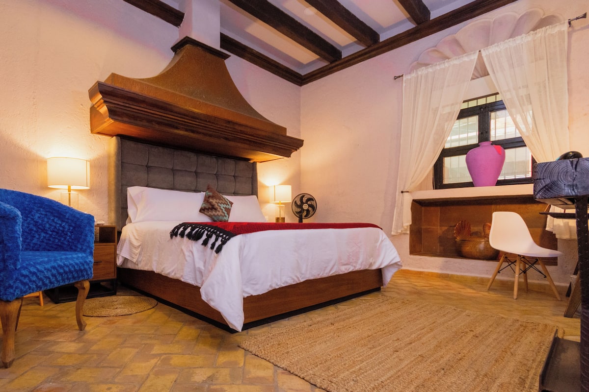 A bedroom with a bed, a blue chair, a wooden nightstand, a fireplace, and a window with curtains.