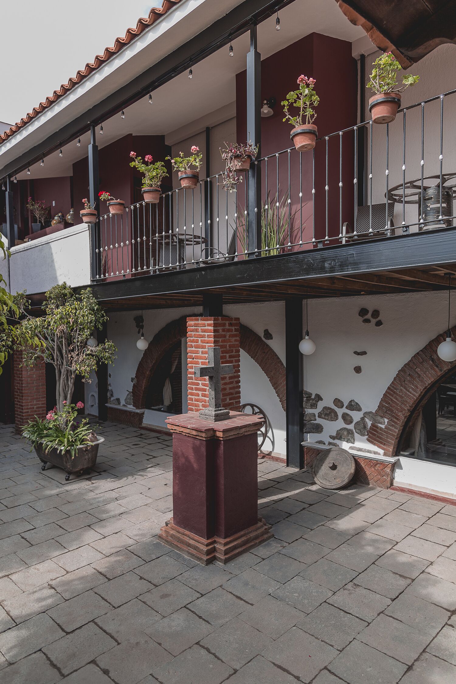 A courtyard with a brick floor, a balcony with potted plants, and a stone wall with a cross.