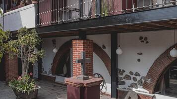 Un patio con suelo de ladrillo, un balcón con macetas y una pared de piedra con una cruz.