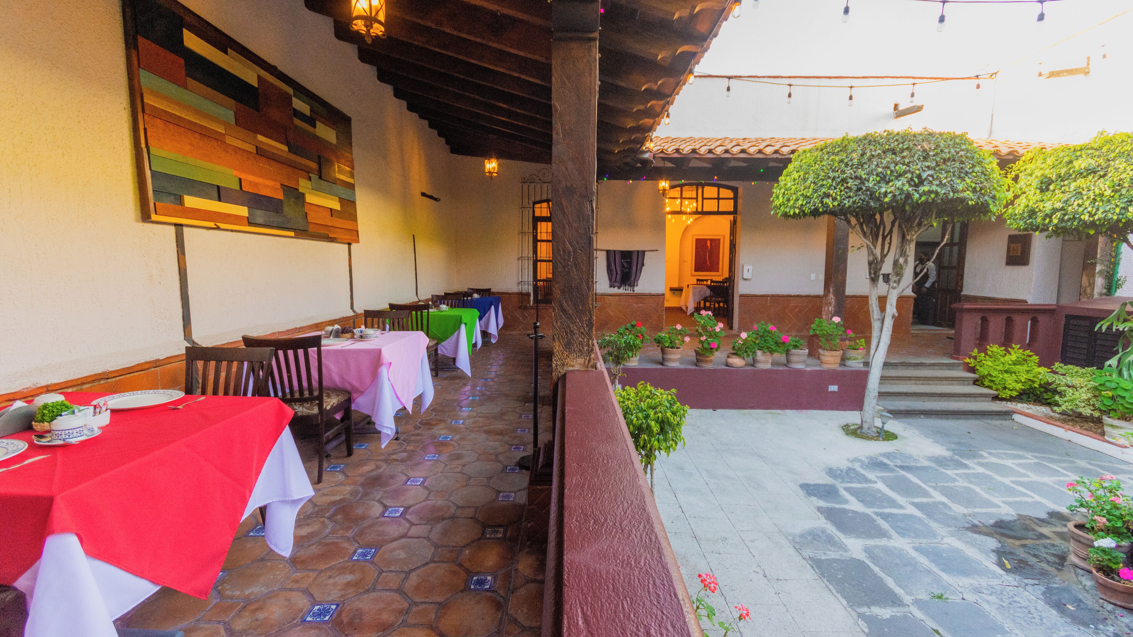 Un área de comedor al aire libre con mesas dispuestas para dos, piso de baldosas de piedra y un pequeño jardín con plantas en macetas y un árbol.