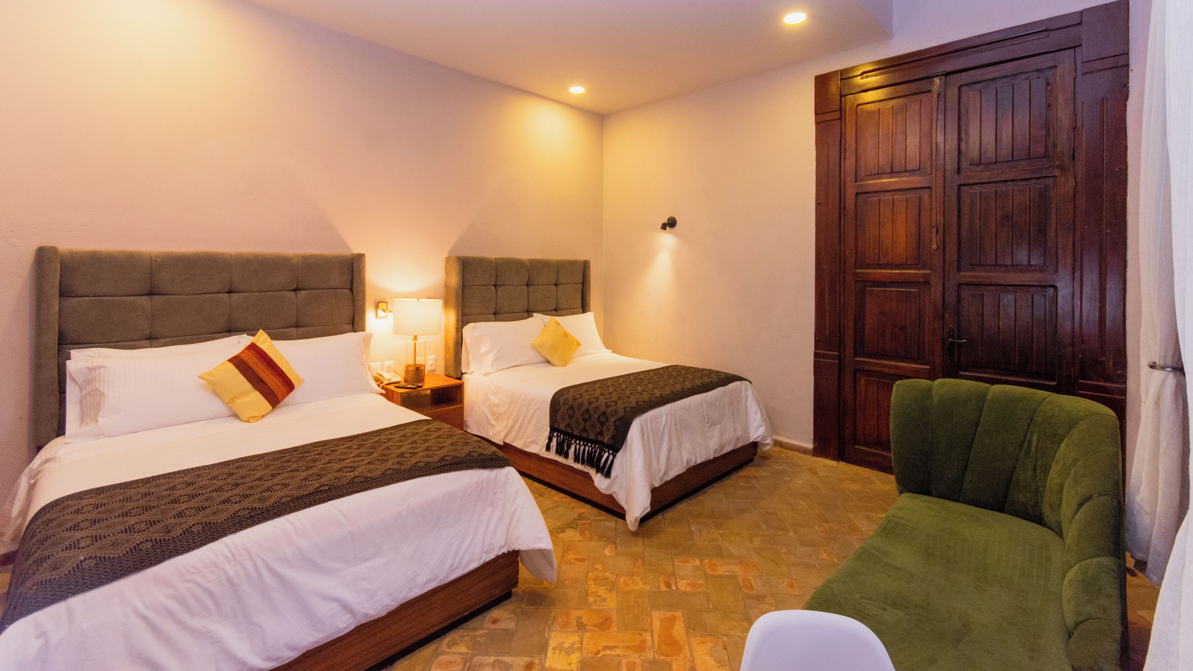 A hotel room with two beds, a green armchair, a wooden door, and a tiled floor.