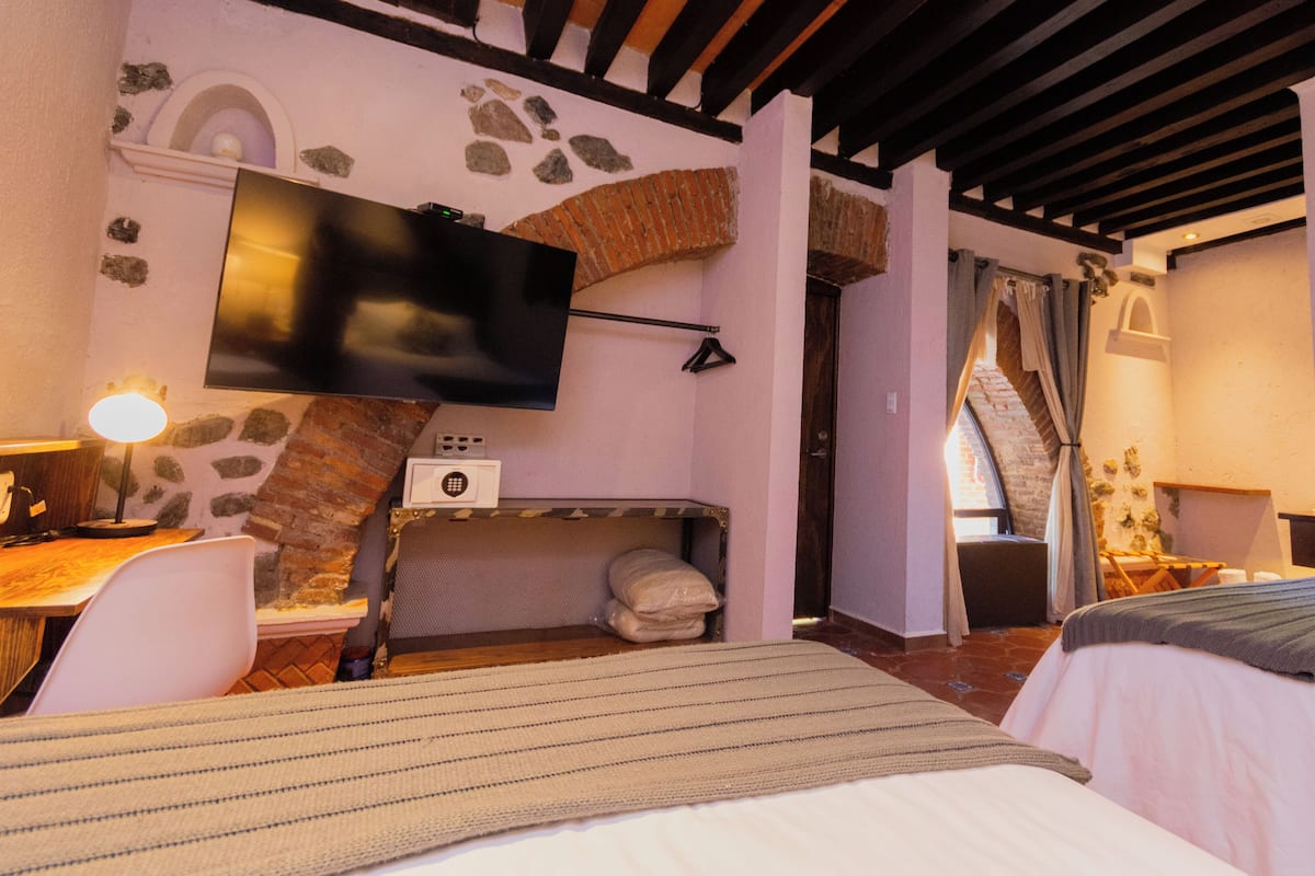 A hotel room with a bed, a desk with a lamp, a TV mounted on the wall, and a stone wall.