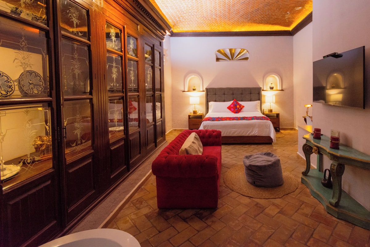 A hotel room with a bed, two bedside tables with lamps, a red sofa, a small round stool, a wooden cabinet with glass doors, and a television mounted on the wall.