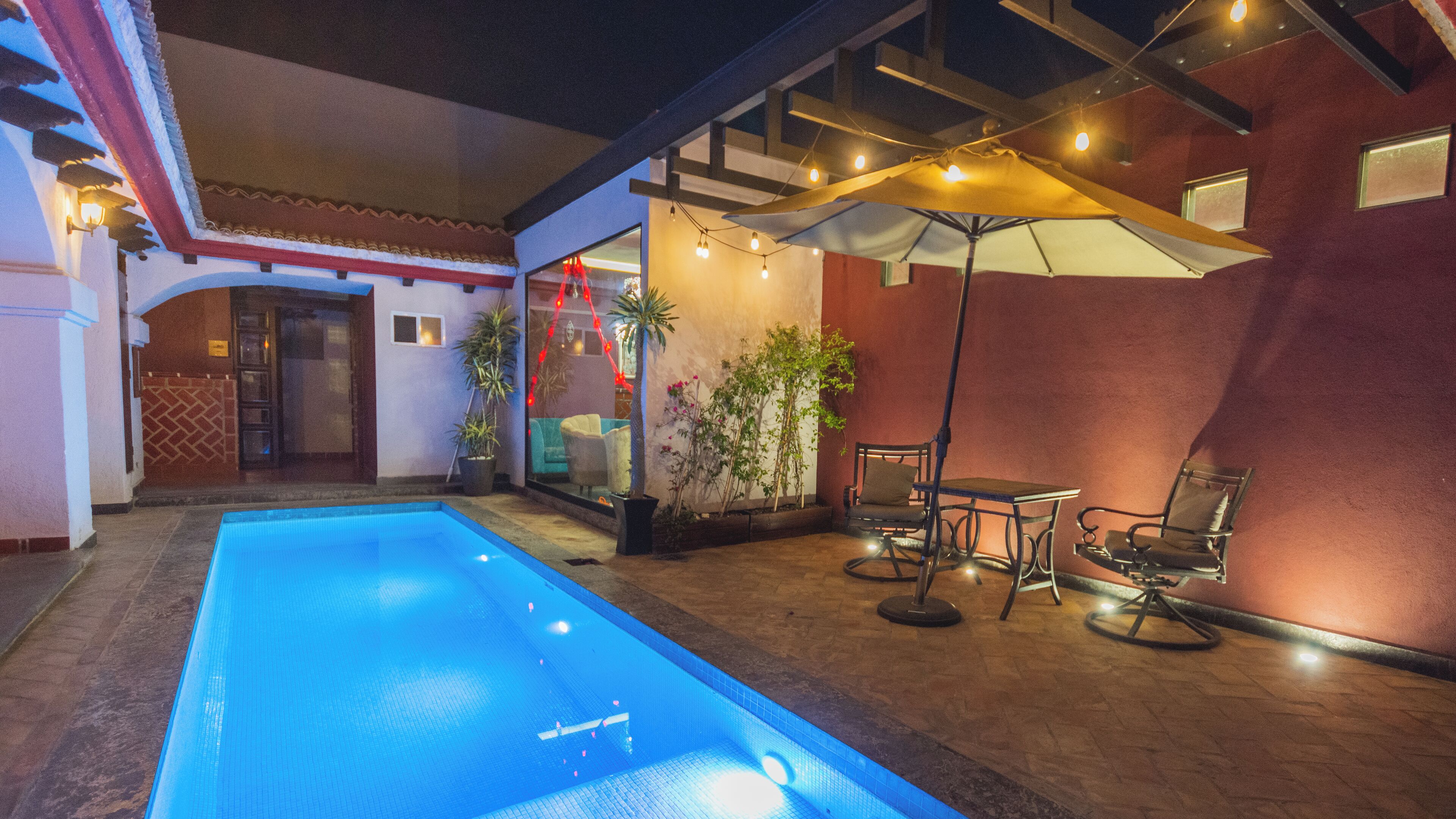 An indoor swimming pool with a tiled floor, surrounded by a patio area with chairs and tables, and a covered seating area with string lights.