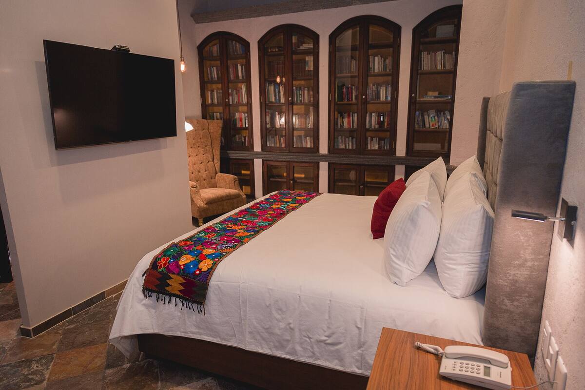 A bedroom with a bed, a bedside phone, a chair, and a TV. There are bookshelves in the background.