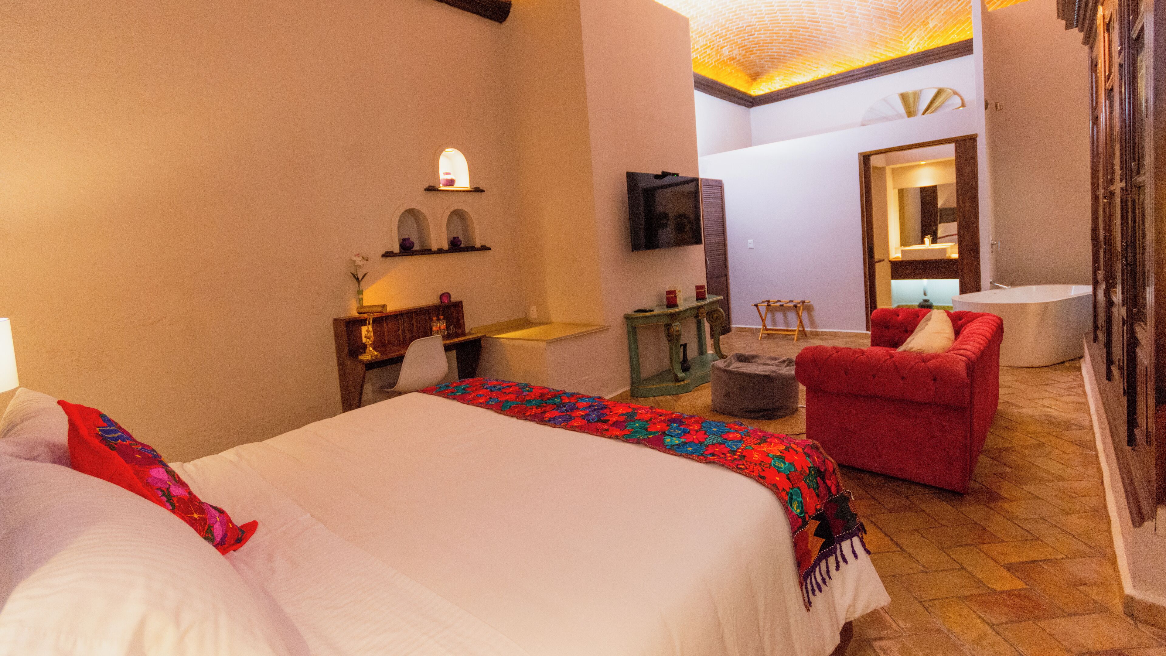 Una habitación de hotel con una cama, un sillón rojo, una mesita, un espejo y un televisor.