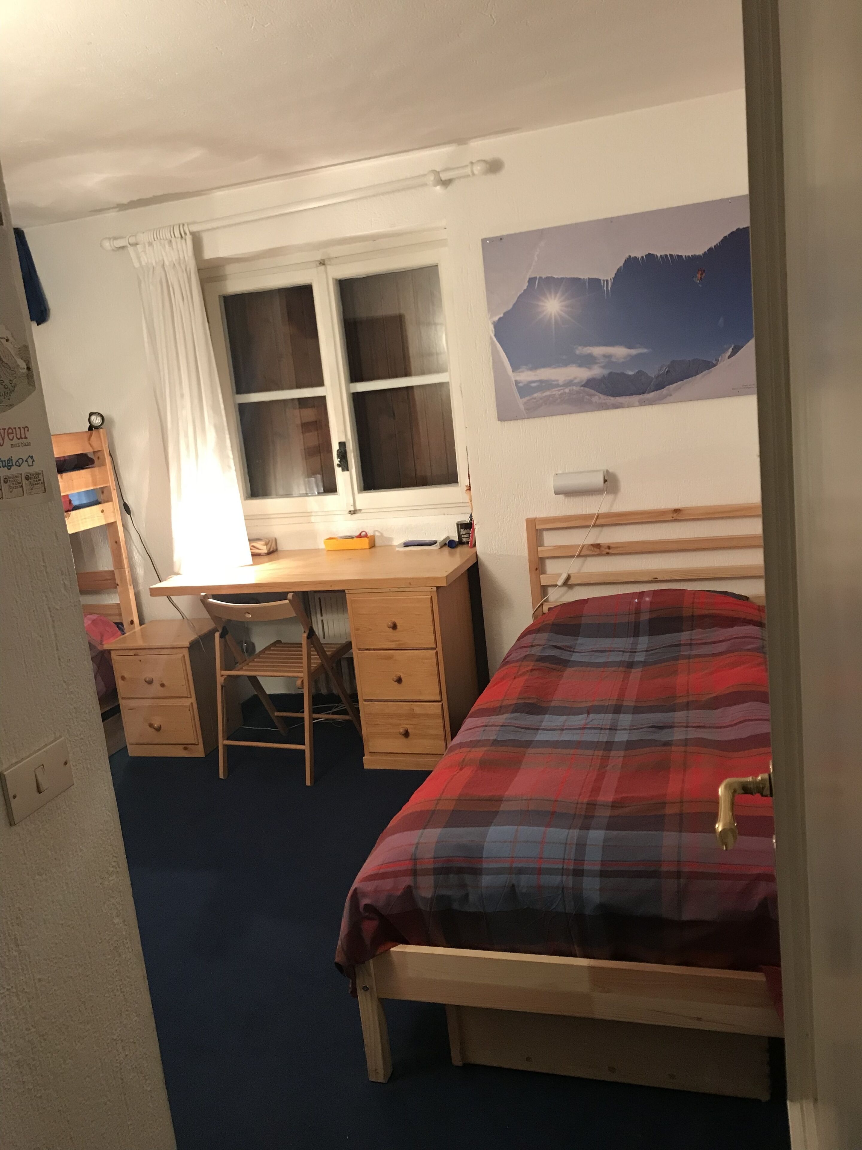A bedroom with a bed, a desk, and a chair. There is a window with curtains, a poster on the wall, and a nightlight on the desk.