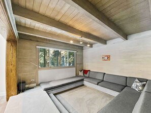 Een modern interieur met een grijze hoekbank, een groot raam met uitzicht op bomen en een houten plafond.