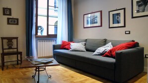 A living room with a grey sofa, red and patterned pillows, a wooden chair, and a small table with books.