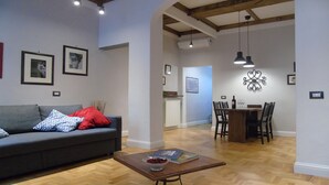 A living room with a grey sofa, red and blue pillows, a wooden coffee table with a bowl and magazines, and a dining area with a table and chairs.